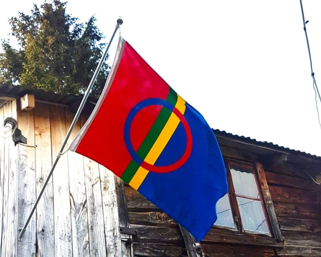 Samisk flagg som henger i en stang på en gammel husvegg. Foto