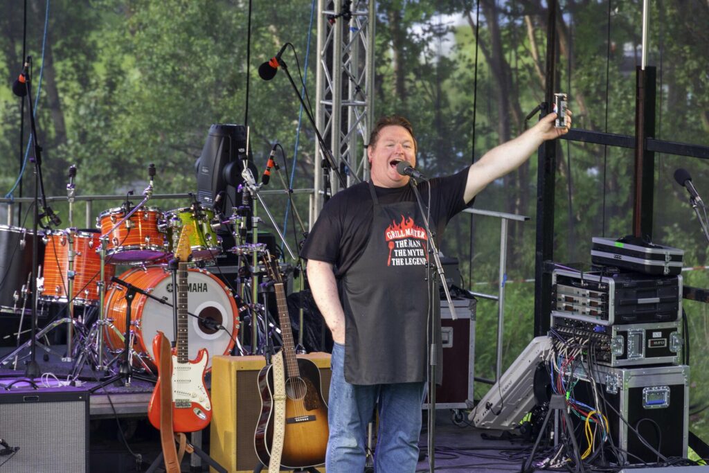 Bilde av mann med forkle som står på scenen med en brusboks i handa mens han snakker i mikrofonen. Foto.