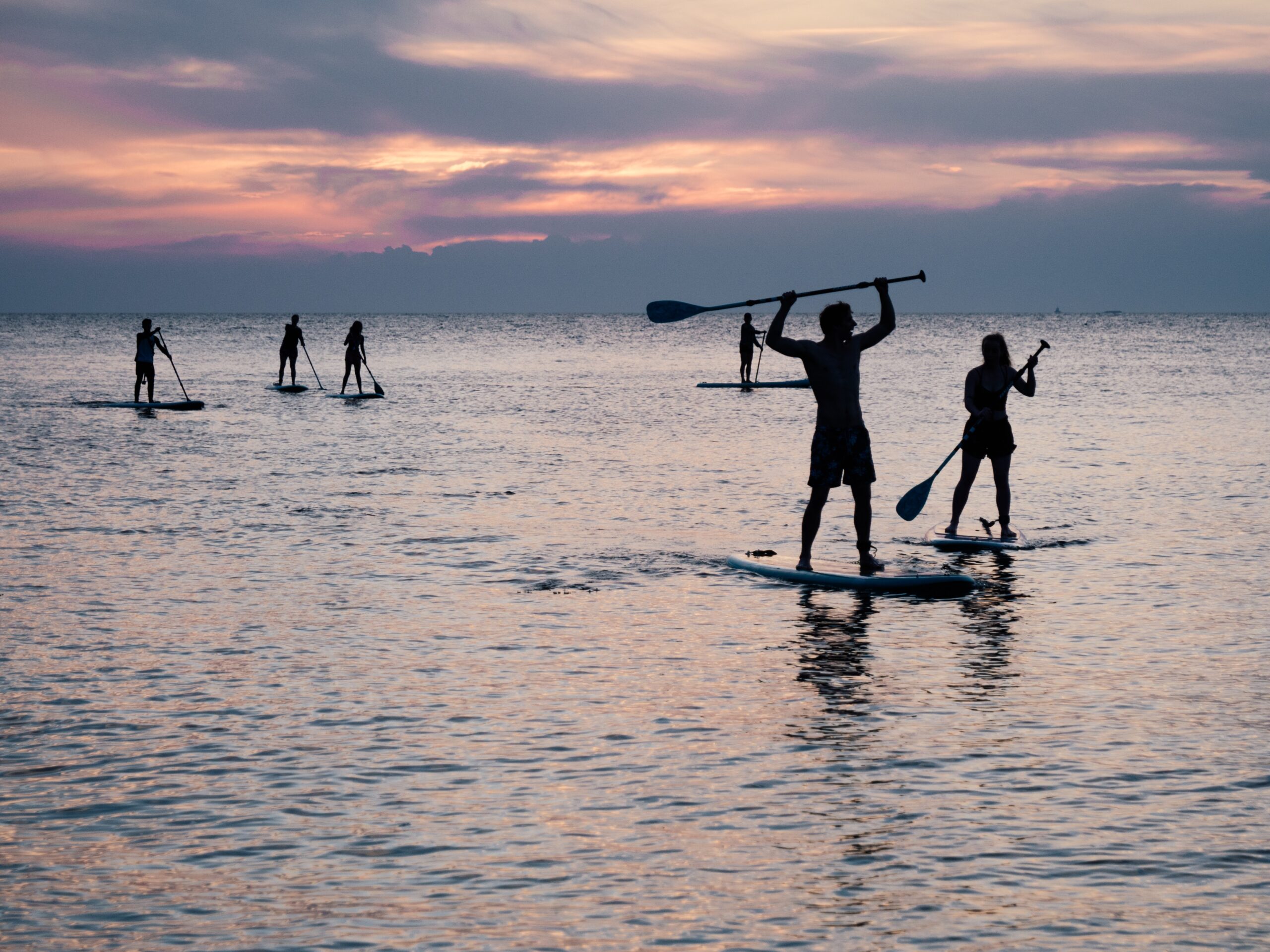 Bilde av flere mennesker som står på surfebrett og padler i solnedgangen. foto