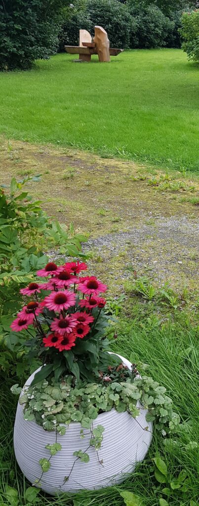 Bilde av en gråhvit vase med rosa blomster framfor en gressplen med en benk som er utskjært av tre i bakgrunnen. foto