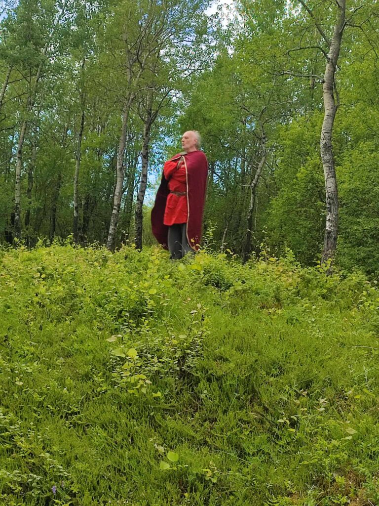 Mann kledd i vikingedrakt står med armene i kors i skogen.foto