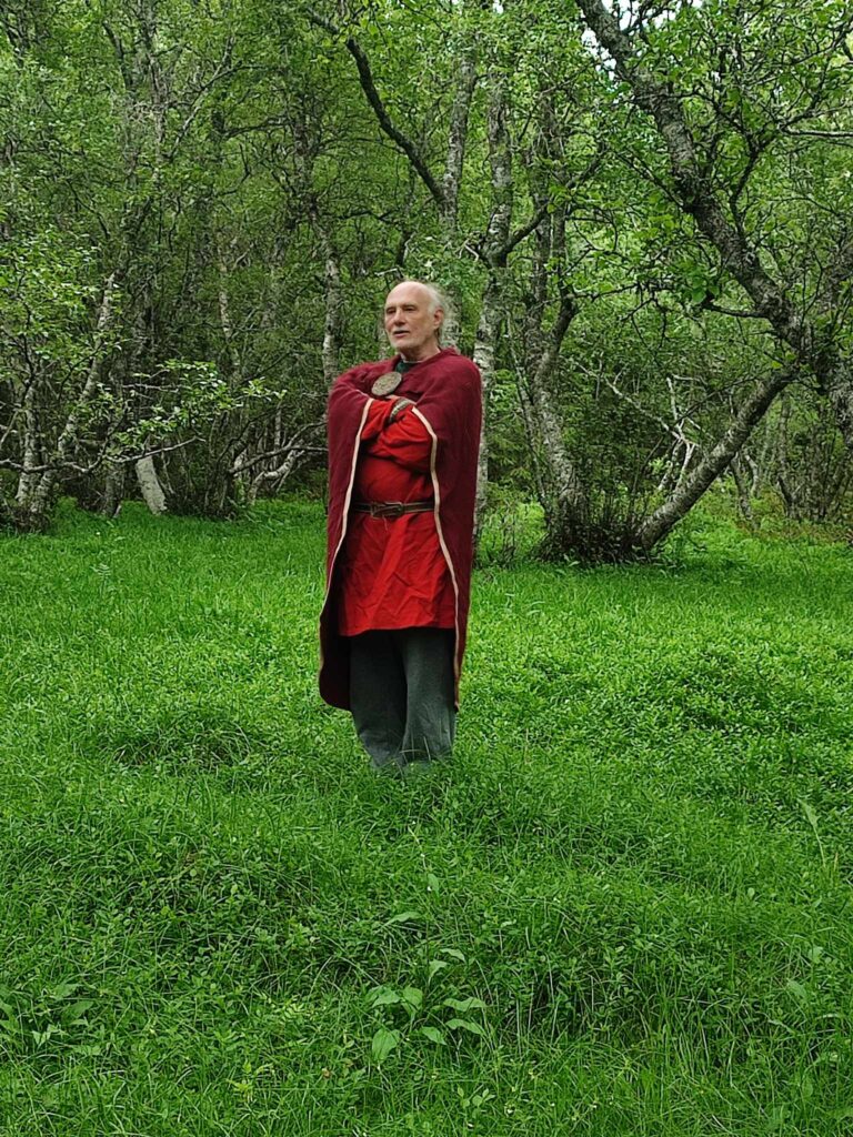 Mann kledd i vikingedrakt står med armene i kors i skogen.foto