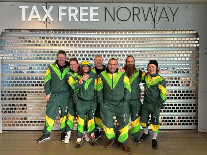 Bandbilde av Mojoo. Bandet står oppstilt foran en stengt tax free-butikk på Bodø Lufthavn. Alle i bandet har på seg like grilldresser med bandets logo på.foto