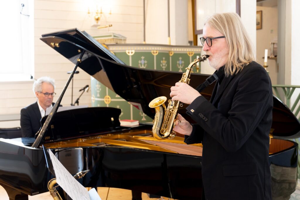 Bilde av mann som spiller sopransaksofon ved siden av et flygel i en kirke. foto