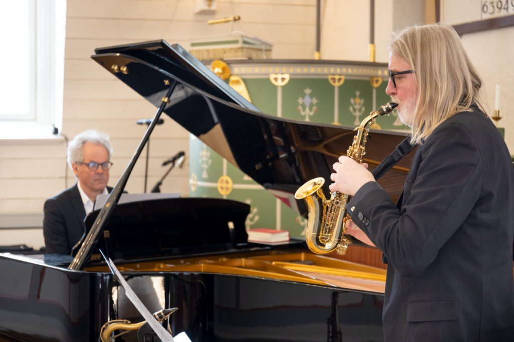 Bilde av mann sett fra siden som står ved et flygel og spiller sopransaksofon. Ved flygelet sitter pianist Jan Gunnar Hoff. foto